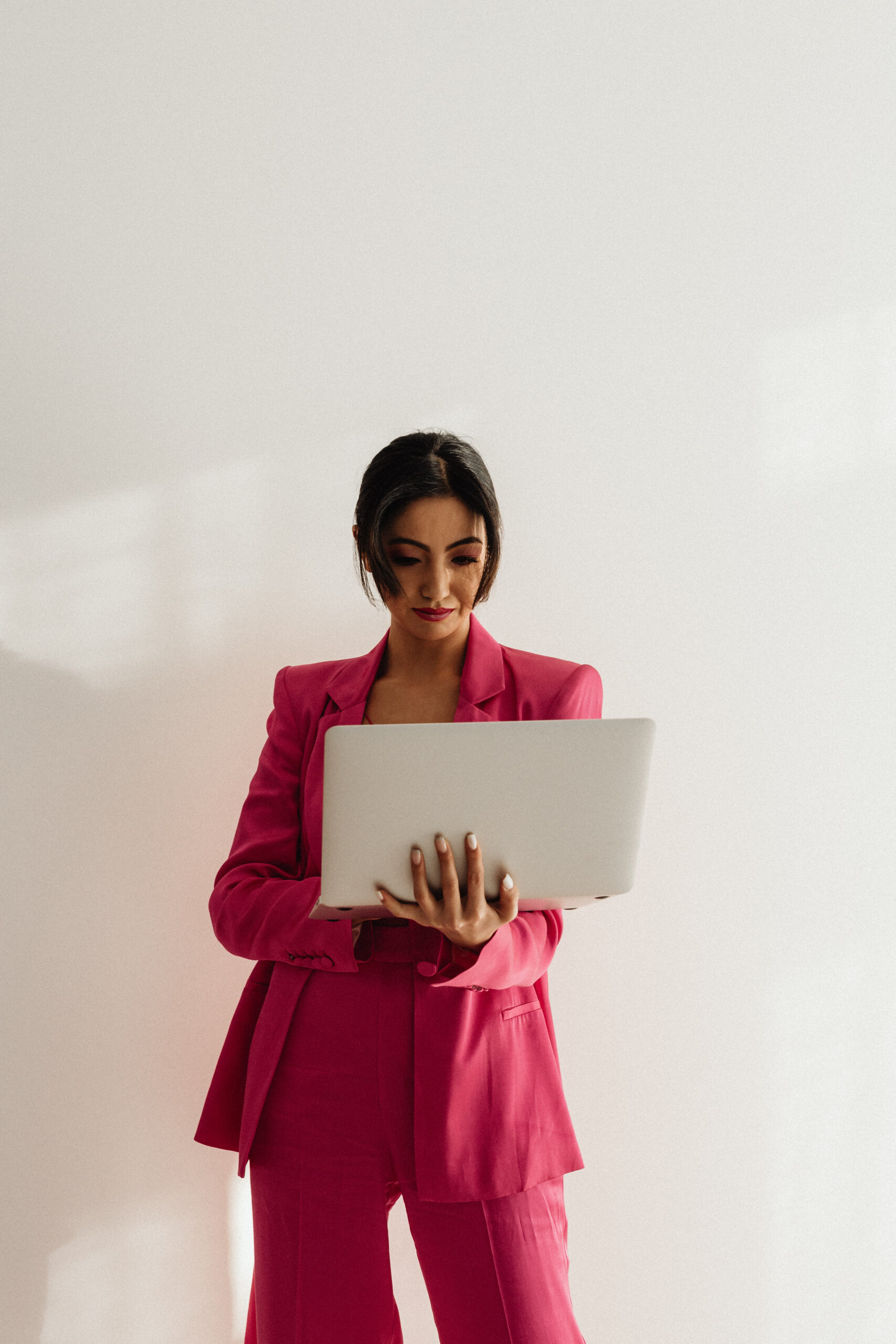 Woman with a pink business suit is looking at a laptop screen that she's holding in her arms.